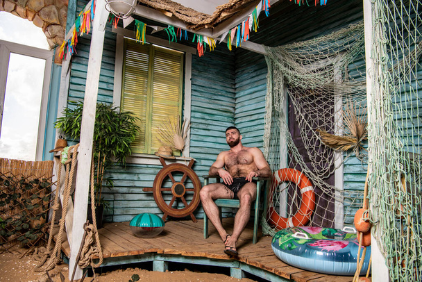 apuesto hombre físicamente bien desarrollado en el porche de una casa de pesca cerca del mar - Foto, Imagen
