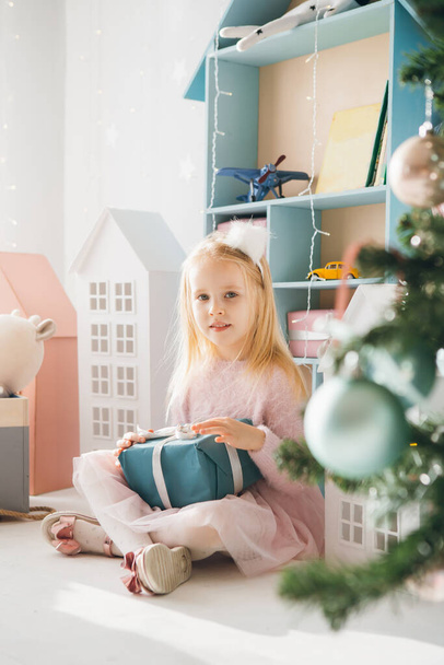 Pretty girl is sitting at the Christmas tree and holding a box with a gift. The holiday concept. Toning. - Foto, imagen