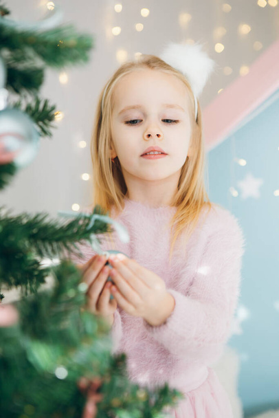 Χαριτωμένο κοριτσάκι με ξανθά μαλλιά στολίζει ένα χριστουγεννιάτικο δέντρο. Η ιδέα των διακοπών. Τόνοι. - Φωτογραφία, εικόνα