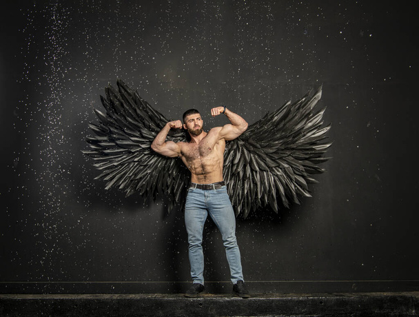 Όμορφος σωματικά ανεπτυγμένος άντρας με μαύρα φτερά αγγέλου σε σκούρο φόντο. - Φωτογραφία, εικόνα