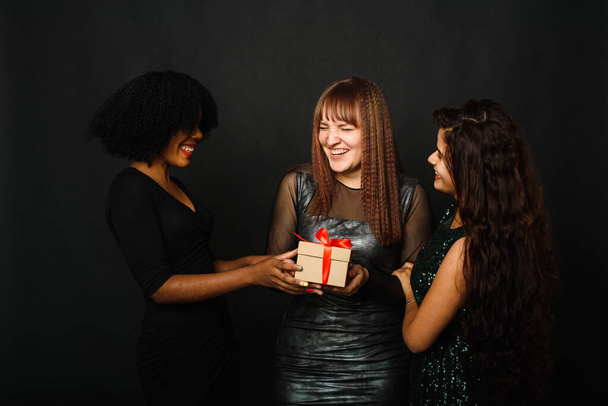 Image of beautiful three young woman rejoicing while standing with present box isolated over black background during christmas celebration - Foto, imagen