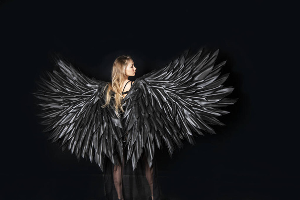 mooi blond meisje in een zwart pak met zwarte vleugels op een zwarte achtergrond - Foto, afbeelding