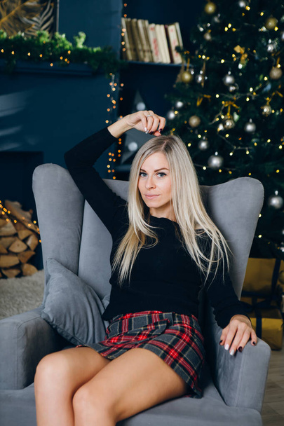 Pretty young woman with blond hair is sitting on a chair against the background of a Christmas tree. New year's concept. Toning. - Valokuva, kuva