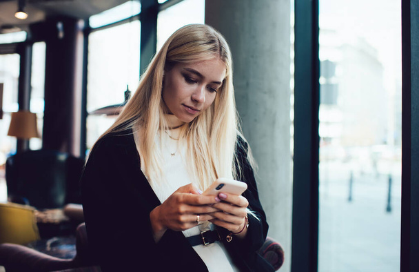 Earnest youthful female with blond hair in casual outfit browsing Internet on mobile phone while standing next to window in cafe - Фото, изображение