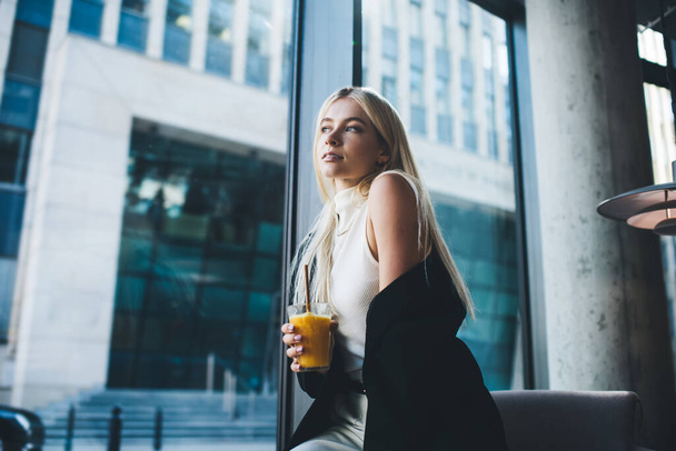 Side view of thoughtful female wearing suit with bare shoulders and drinking fresh juice while spending time in modern restaurant - Photo, image