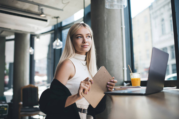 Young businesswoman with blond hair holding clipboard in hands and looking away while working with papers and using laptop in cafe - Foto, afbeelding