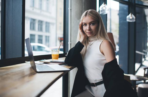 Young elegantly dressed female with blond hair looking at camera while working with documents and using laptop in modern cafe - 写真・画像
