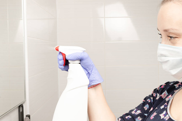 roztomilý mladá dívka služka nosí lékařskou masku sype čisticí prostředek na zrcadle v koupelně během pandemie coronavirus - Fotografie, Obrázek