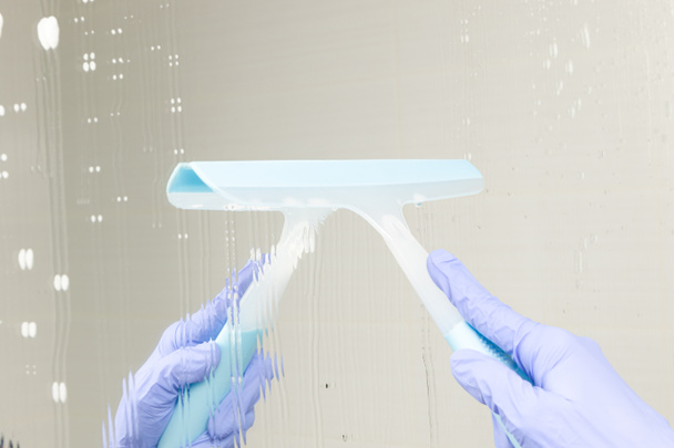 женская рука в синей резиновой перчатке крупным планом вытирает зеркало в ванной комнате на фоне белой плитки - Фото, изображение