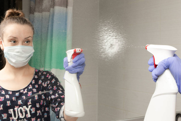 χαριτωμένο νεαρή κοπέλα υπηρέτρια φορώντας μια ιατρική μάσκα ψεκάζει καθαριστικό παράγοντα στον καθρέφτη του μπάνιου κατά τη διάρκεια της πανδημίας coronavirus - Φωτογραφία, εικόνα