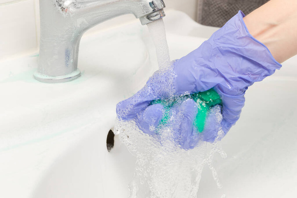θηλυκό χέρι σε ένα μπλε γάντι πλένει ένα πράσινο σφουγγάρι καουτσούκ αφρού κάτω από μια βρύση στο μπάνιο. Καθαρισμός σπιτιού - Φωτογραφία, εικόνα
