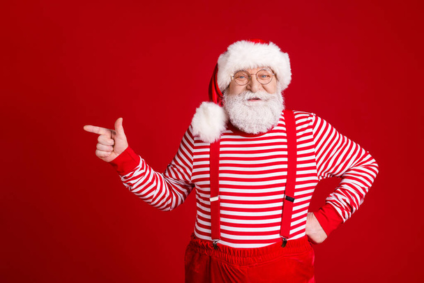 Photo of pensioner old man direct finger empty space suggest fine christmas clothes shop wear santa costume suspenders spectacles striped shirt headwear isolated red color background - Valokuva, kuva