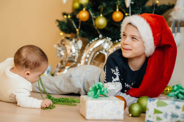 A little boy and his brother play with gifts under a festive Christmas tree. Happy Christmas and New Year celebrations - Foto, Imagen