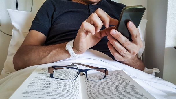 Vista da vicino del paziente di sesso maschile in recupero dopo un intervento chirurgico utilizza lo smartphone mentre è sdraiato su un letto in ospedale con gli occhiali appoggiati su un libro - Foto, immagini