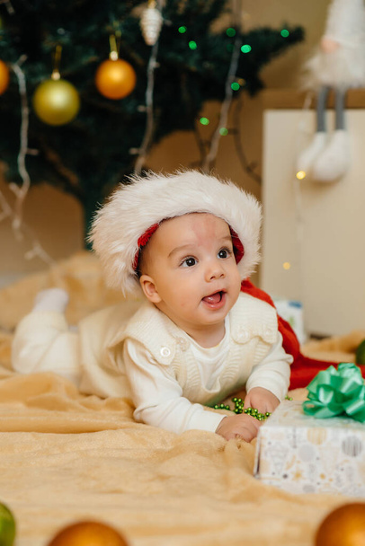 Cute smiling baby is lying under a festive Christmas tree and playing with gifts. Christmas and New Year celebrations - Foto, Bild