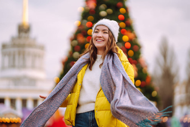 Happy woman on Christmas market. Young woman in winter style clothes posing at festive street market. Lights around. New year tree. - Фото, зображення