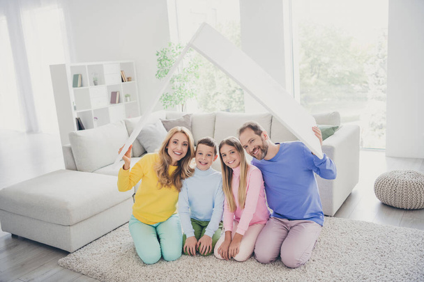Full body photo of family mommy daddy sit floor carpet hold paper card roof above little kids boy girl in house indoors - Foto, Bild