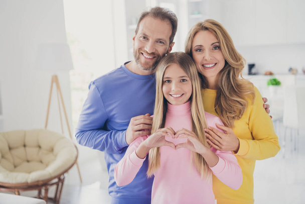 Photo of positive cheerful family mom dad stand behind small kid girl show heart finger sign in house indoors - Valokuva, kuva