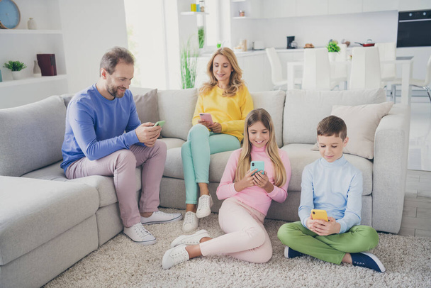 Full length photo of mommy daddy and two little kids texting on smartphone sit sofa floor in house indoors - 写真・画像
