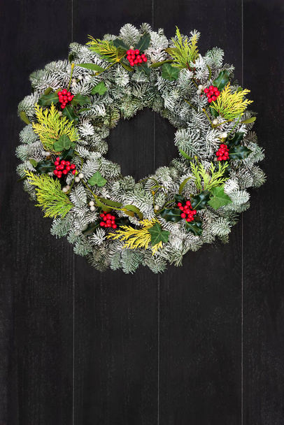 冬&雪で覆われたスプルースモミ、杉、ホリー、ミステリー&ツタの葉でクリスマスリース素朴なダークウッドのフロントドアの背景にあります。クリスマスの休日の季節と新年のための作曲. - 写真・画像