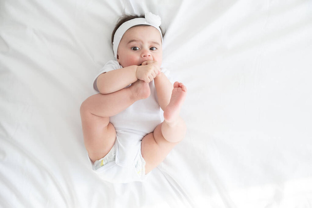 funny 6 months baby girl playing with legs, lying on bed at home on white bedding. top view. - Fotografie, Obrázek