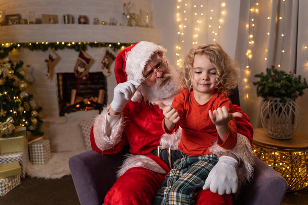 Funny Santa Claus sitting on his rocker with little cute boy sitting on his knee. Christmas spirit, holidays and celebrations concept - Foto, imagen