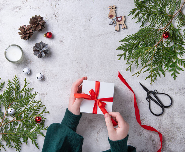Filles ouverture blanc cadeau de Noël avec ruban rouge sur table gris clair. Fond de Noël avec thuja arbre, bougies cônes et jouets de Noël. Couché plat et vue du dessus - Photo, image