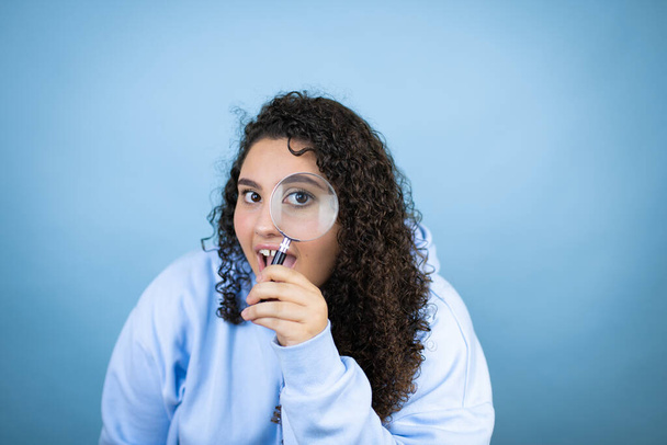 Mujer hermosa joven con sudadera casual sobre fondo azul aislado sorprendida mirando a través de una lupa - Foto, Imagen