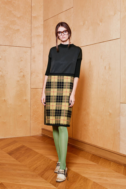 Full length portrait of a pretty brunette girl wearing a black shirt and plaid skirt and in green tights. Standing pose on plywood background. - Foto, afbeelding