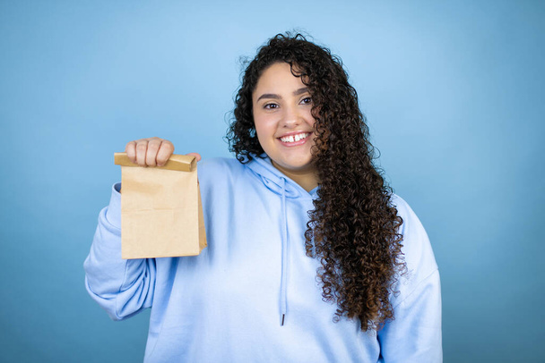 Junge schöne Frau trägt lässiges Sweatshirt über isoliertem blauem Hintergrund, lächelt und hält eine Papiertüte in der Hand - Foto, Bild