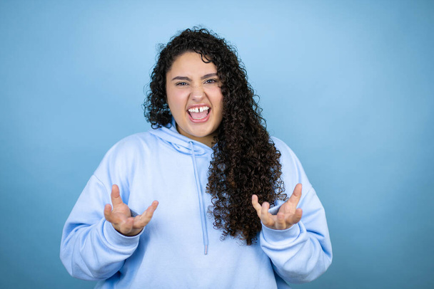 Young beautiful woman wearing casual sweatshirt over isolated blue background crazy and mad shouting and yelling with aggressive expression and arms raised. Frustration concept. - Photo, Image