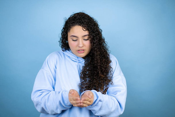 Junge schöne Frau in lässigem Sweatshirt über isoliertem blauem Hintergrund, lächelnd mit den Handflächen, die Geste empfangend oder gebend. Halt und Schutz - Foto, Bild