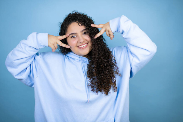 Young beautiful woman wearing casual sweatshirt over isolated blue background Doing peace symbol with fingers over face, smiling cheerful showing victory - Φωτογραφία, εικόνα