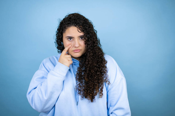 Junge schöne Frau in lässigem Sweatshirt über isoliertem blauem Hintergrund Zeigt auf das Auge, das dich beobachtet, Geste, misstrauischer Ausdruck - Foto, Bild