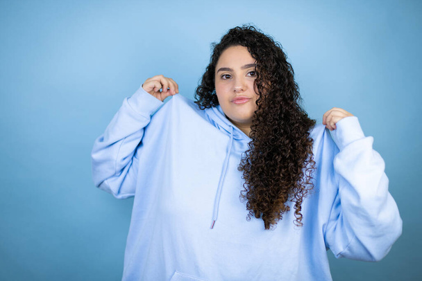 İzole edilmiş mavi arka plan üzerine sıradan bir sweatshirt giyen genç ve güzel bir kadın tişörtünü başarılı bir ifadeyle tutuyor. - Fotoğraf, Görsel