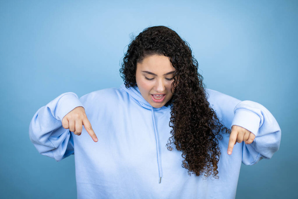 Junge schöne Frau in lässigem Sweatshirt vor isoliertem blauen Hintergrund überrascht, schaut nach unten und zeigt mit erhobenen Armen und Fingern nach unten - Foto, Bild