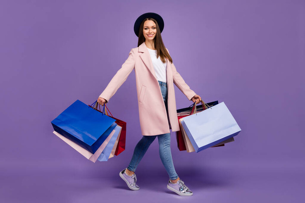 Full length profile side photo of cheerful lovely trendy girl hold many bags go from shopping mall wear beige outfit denim headwear sneakers isolated over purple color background - 写真・画像