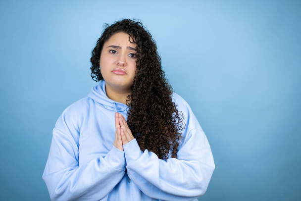 İzole edilmiş mavi arka plan üzerine sıradan bir sweatshirt giyen genç ve güzel bir kadın elleri bağlı yalvarıyor ve dua ediyor. Yüzünde umut ifadesi var. Çok duygusal ve endişeli. - Fotoğraf, Görsel