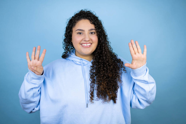 Молода красива жінка носить повсякденний светр над ізольованим синім тлом, показуючи і вказуючи пальцями номер дев'ять, посміхаючись впевнено і щасливо
 - Фото, зображення