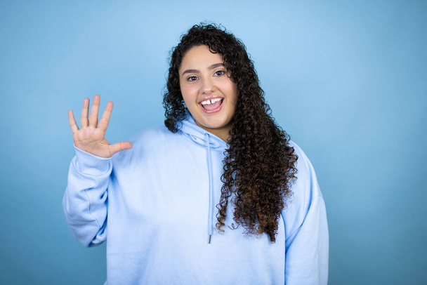 İzole edilmiş mavi arka plan üzerine sıradan bir sweatshirt giyen genç ve güzel bir kadın beş numaralı parmaklarıyla işaret ederken kendinden emin ve mutlu bir şekilde gülümsüyor. - Fotoğraf, Görsel