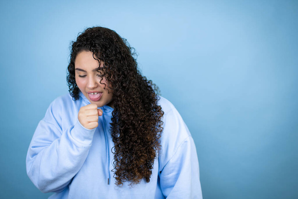 Νεαρή όμορφη γυναίκα που φοράει ένα απλό φούτερ πάνω από απομονωμένο μπλε φόντο με το χέρι της στο στόμα της επειδή βήχει - Φωτογραφία, εικόνα