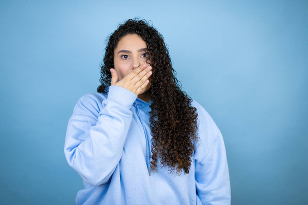 Junge schöne Frau in lässigem Sweatshirt über isoliertem blauem Hintergrund überrascht, den Mund zu bedecken - Foto, Bild