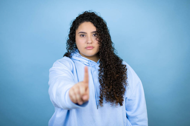 Jonge mooie vrouw dragen casual sweatshirt over geïsoleerde blauwe achtergrond tonen en wijzen met vingers nummer een terwijl is serieus - Foto, afbeelding