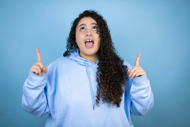 Junge schöne Frau in lässigem Sweatshirt vor isoliertem blauen Hintergrund erstaunt und überrascht in die Kamera blickend und mit erhobenen Fingern und Armen nach oben zeigend - Foto, Bild