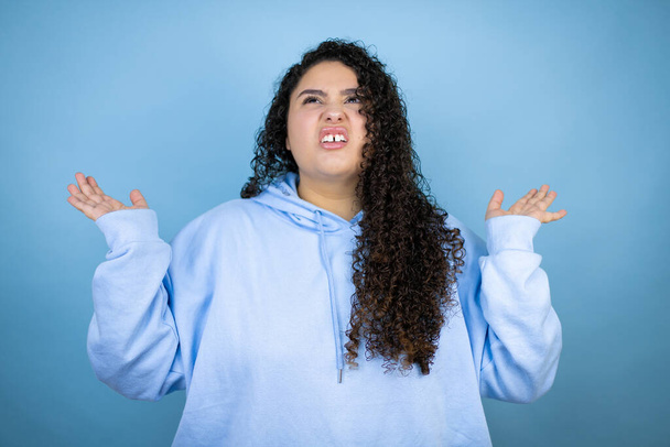 Jonge mooie vrouw dragen casual sweatshirt over geïsoleerde blauwe achtergrond clueless en verwarde uitdrukking met armen en handen opgeheven - Foto, afbeelding