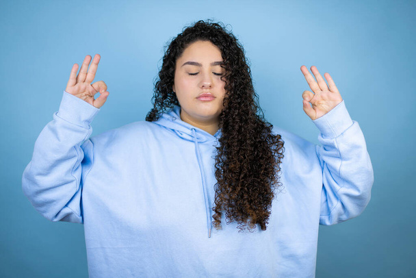 Junge schöne Frau trägt lässiges Sweatshirt über isoliertem blauem Hintergrund und lächelt mit geschlossenen Augen und macht Meditationsgeste mit den Fingern - Foto, Bild