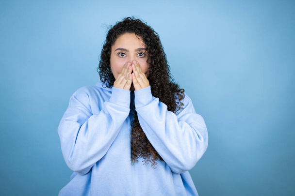 Young beautiful woman wearing casual sweatshirt over isolated blue background with her hands over her mouth and surprised - Photo, Image