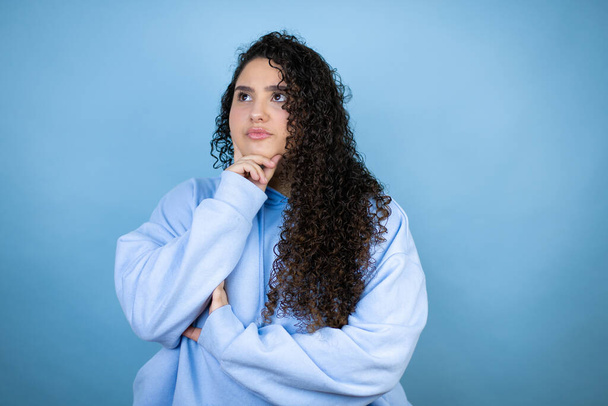 Fiatal gyönyörű nő visel alkalmi pulóver felett elszigetelt kék háttér gondolkodás és látszó oldalra - Fotó, kép