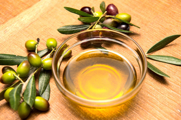 Extra virgin olive oil in a clear bowl on a wooden background and a bowl of olives and olive branches - Фото, изображение