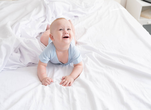 Смешной мальчик улыбается и лежит на белом постельном белье дома.  - Фото, изображение